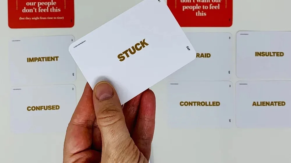 Mână ținând un card cu emoția „STUCK”, în fața unui perete cu carduri de emoții nedorite: impatient, confused, controlled, alienated.