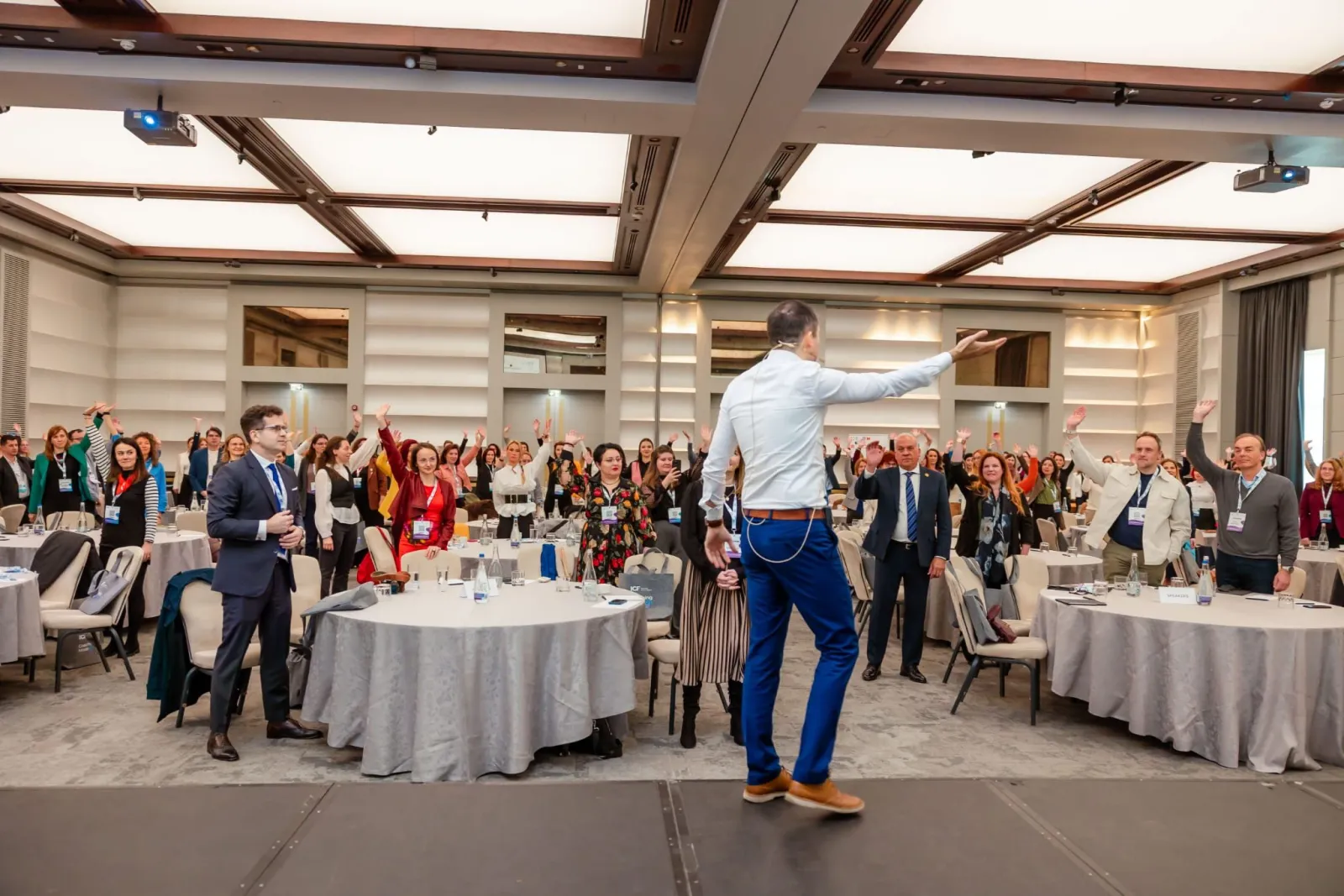 A moderator at the edge of the stage, arm outstretched to the room, opening a conference as hundreds of attendees raise their hands.